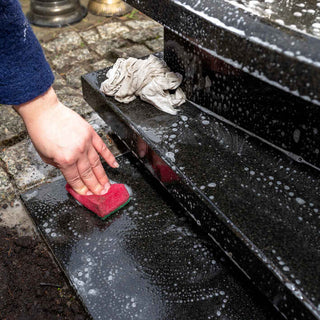 person scrubbing a black tombstone with a red sponge and soapy water