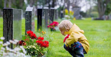 little boy in a yellow rain coat crouching in front of a headstone with flowers in front of it